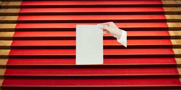 A stairwell with a red and gold carpet is in the background. In the foreground a floating hand is a holding a book.