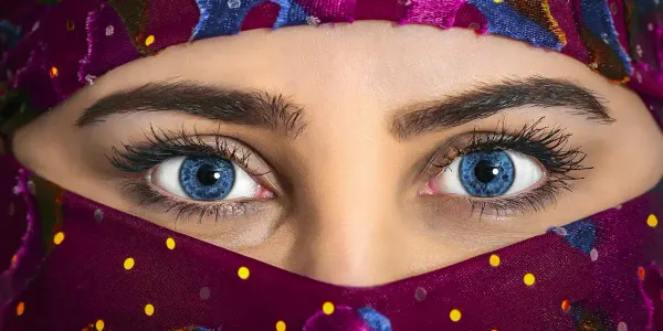A close-up shot of a woman's face. Her bold blue eyes are looking straight at the viewer in a piercing way. The rest of her face is covered by a hijab.