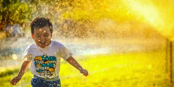 A smiling child is running through a spray of water from a sprinkler on a sunny day